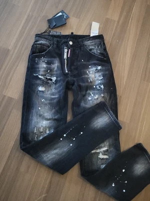 Dsq2 jeans  - Säljer ett par svarta jeans med coola slitningar och vita färgstänk. Jeansen har raka ben, klassisk femficksmodell och detaljer som ger en edgy streetkänsla. Materialet är denim med tvättad finish och dragkedja fram. Perfekta för dig som gillar en trendig och avslappnad stil.