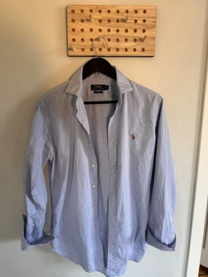 Ljusblå skjorta från Polo Ralph Lauren - Klassisk ljusblå skjorta från Polo Ralph Lauren med broderad logga på bröstet. Skjortan har lång ärm, knappar framtill och en avslappnad passform. Perfekt till jeans eller chinos för en clean och stilren look.