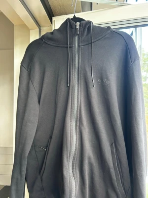 Svart hoodie med dragkedja från BOSS - Säljer en svart hoodie från BOSS med dragkedja framtill och två fickor. Hoodien har justerbar huva med snören och diskret BOSS-logga på bröstet. Tillverkad i mjukt sweatshirtmaterial som är skönt att ha på sig.