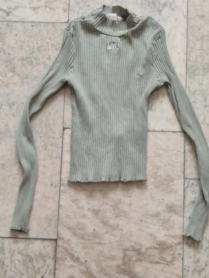 Ribbad grå polotröja med NYC-tryck - Snygg ribbad polotröja med b'NYC' broderat framtill. Grön stretchig ribbstickad tröja med långa ärmar.  Fint skick förutom ett mycket litet hål. (Se bilder)