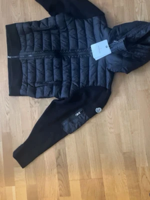 Svart Moncler Cardigan - Snygg svart Cardigan från Moncler med huva och dragkedja framtill. Jackan har quiltad kropp, släta ärmar och en Moncler-logga på ena ärmen. Praktiska fickor med dragkedja och en sportig look som passar perfekt till vintern.