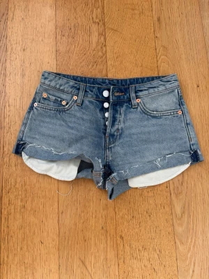 Blå denimshorts med rå kant - Säljer ett par blå jeansshorts från weekday i klassisk femficksmodell med midwaist och rå, fransig kant. Shortsen har knäppning framtill med tre synliga knappar och uppvikta ben. Perfekta för sommaren och ger en avslappnad look.