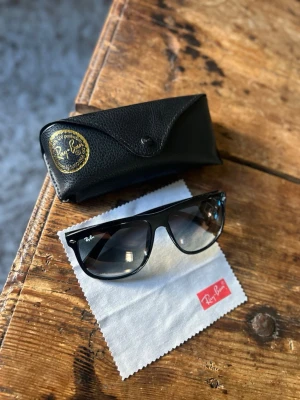 Svarta Ray-Ban boyfriend’s solglasögon - Klassiska svarta Ray-Ban boyfriend’s solglasögon med blank finish och mörka glas. Säljs med originalfodral i svart skinn och putsduk med Ray-Ban-logga. Perfekta för dig som vill ha en tidlös och ikonisk look.