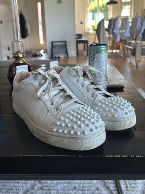 Vita Louboutin sneakers med nitar - Säljer ett par vita sneakers från Christian Louboutin med ikoniska röda sulor och coola nitar på tån. Skorna har lågt skaft, vita snören och är tillverkade i läder. Perfekta för dig som vill sticka ut med en edgy och lyxig stil. OBS, ingen box följs me, EN NIT SAKNAS PÅ VÄNSTER SKON.