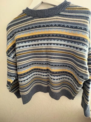 Mönstrad stickad tröja Pull&Bear XS - Stickad tröja från Pull&Bear i storlek XS med grå, blå, vit och gul mönstrad design. Tröjan har rund halsringning och ribbade muddar. Perfekt för dig som gillar retrovibbar och mysiga plagg.