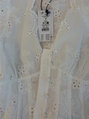 Vit broderad blus med knappar(oanvänd) - Romantisk vit blus med broderade blommor och blad, v-ringad med vågig kant och klädda knappar framtill. Långärmad modell med lätt puff vid axlarna och figurnära passform. Perfekt till jeans eller kjol för en trendig look. Säljer pågrund av att den är för stor!