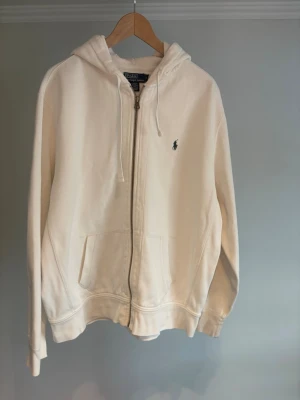  Polo Ralph Lauren zip hoodie  - Klassisk vit hoodie från Polo Ralph Lauren med dragkedja framtill och två fickor. Har huva med dragsko och den ikoniska lilla broderade loggan på bröstet. Tillverkad i mjukt bomullsmaterial som är perfekt för chill dagar. Nypris runt 2500 kr. Personen på bild är 175 cm 
