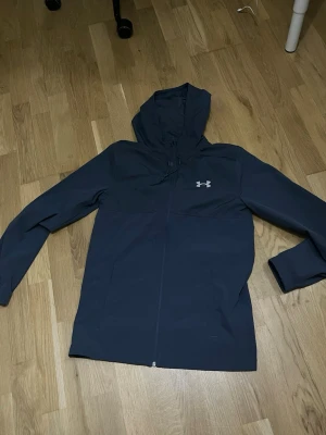 Mörkblå vindjacka från Under Armour - Snygg mörkblå vindjacka från Under Armour med huva och dragkedja framtill. Jackan har logga på bröstet och stor text på ryggen. Tillverkad i ett lätt och vindtätt material, perfekt för blåsiga dagar.