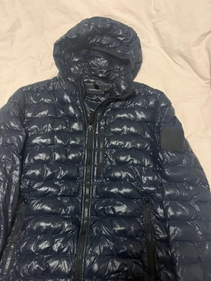 Mörkblå pufferjacka Peak Performance - Snygg mörkblå pufferjacka från Peak Performance med huva och flera dragkedjeförsedda fickor. Jackan har quiltad design och glansigt yttertyg. Perfekt höst jacka som är i jättebra skick.