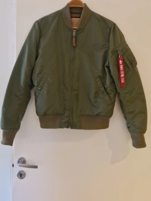 Alpha Industries Olivgrön bomberjacka med dragkedja - Äkta Aplha Industries jacka i snygg olivgrön färg med ribbade muddar och krage. Jackan har en klassisk armficka med röd tag och dragkedja framtill, två sidofickor med knappstängning och en innerficka. Tillverkad i glansigt material som ger en cool look. Perfekt för dig som gillar streetwear och tidlös stil.