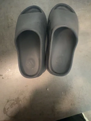 Svarta Yeezy slides - Säljer ett par svarta slides tofflor från Adidas med bred rem över foten och öppen tå. Tillverkade i ett mjukt och flexibelt syntetmaterial som ger skön komfort. Perfekta för chill dagar hemma eller till stranden.