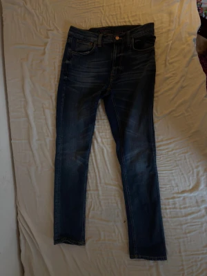 Mörkblå smala nudie jeans - Storlek W31 L30 Snygga mörkblå jeans med smal passform och klassiska fem fickor. Jeansen har subtila slitningar och kontrastsömmar för en trendig look. Tillverkade i denim med stretch för extra komfort. Perfekta till vardagsstilen.