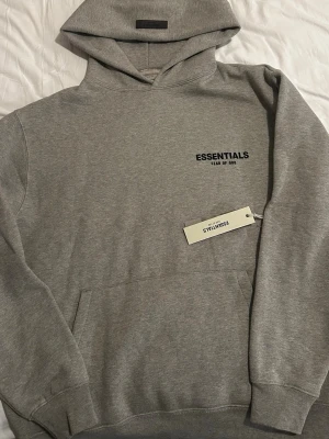 Grå hoodie Essentials Fear of God - Säljer en stilren grå hoodie från Essentials Fear of God. Hoodien har huva, magficka och svart logotyptryck på bröstet. Tillverkad i mjukt bomullsmaterial som är perfekt för en avslappnad look. Passar dig som gillar streetwear och enkel design.
