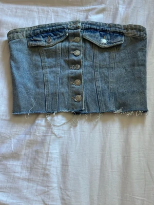 Blå ”jeans” tubtopp med knappar - Säljer en blå denim tubtopp med silverfärgade knappar framtill och två dekorativa bröstfickor. Toppen har rå, fransig kant nedtill och smockad rygg för skön passform. Perfekt för dig som gillar en edgy och trendig stil.