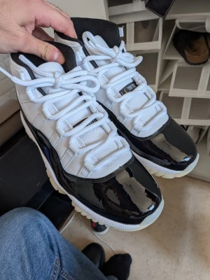 Svarta och vita Air Jordan 11 sneakers - Säljer ett par Air Jordan 11 sneakers i svart och vitt med gulddetaljer. Skorna har blankt lackat läder runt om, vit mesh på ovandelen och klassisk Jumpman-logga på sidan. Tjock vit sula med isblå inslag och högt skaft. Perfekta för dig som gillar ikoniska basketskor. Oanvända.