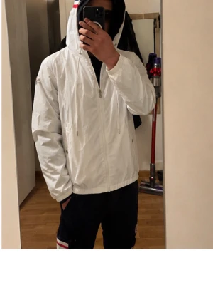 Moncler Windbreaker - Snygg vit vindjacka med dragkedja och huva med dragsko. Jackan har en enkel och clean design med diskret logga på ärmen och fickor med tryckknapp. Perfekt för blåsiga dagar och lätt att matcha med olika outfits.