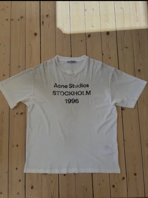 Vit Acne Studios t-shirt med tryck, stl. S - Vit t-shirt från Acne Studios med svart texttryck på bröstet där det står 'Acne Studios STOCKHOLM 1996'. Klassisk rund hals och korta ärmar. Tillverkad i mjuk bomull för en skön och avslappnad stil. Distressed stil. Nypris 3.300 kr. Endast provad. Etikett medföljer 