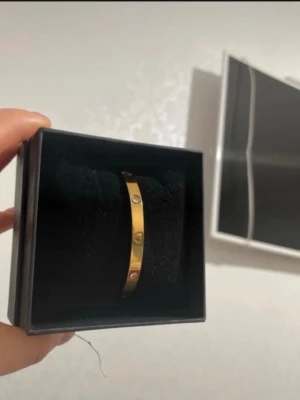 Guldigt armband med hjärtdetaljer - Snyggt armband i guldfärgad metall med infällda stenar och hjärtdetaljer. Armbandet har en stilren och modern design som passar perfekt till din vardagsstil. Levereras i en svart presentask.
