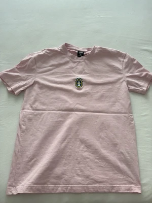 Ljusrosa t-shirt från FSBN - Säljer en ljusrosa t-shirt från FSBN i relaxed fit. T-shirten har korta ärmar, rund halsringning och ett litet broderat emblem på bröstet. Tillverkad i mjuk bomull, perfekt för en chill och enkel stil. Jag köpte den i New York har bara använts den 2 gånger 