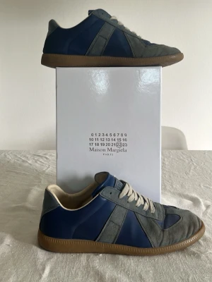 Maison Margiela blå/grå sneakers - Snygga sneakers från Maison Margiela i blått och grått med ovandel i mocka och läder. Skorna har vita snören och en klassisk gummisula. Perfekt för dig som gillar stilrena och trendiga detaljer.