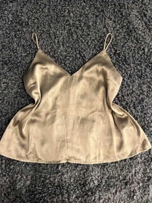Champagnefärgad linnetopp i satin - Säljer en champagnefärgad linnetopp i satin med tunna axelband och v-ringad hals. Toppen har en lätt glansig finish och är perfekt för att lyfta vilken outfit som helst. Enkel och stilren design som känns både trendig och tidlös.