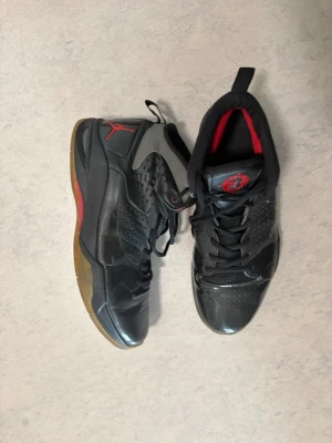Svarta Air Jordan sneakers - Säljer ett par svarta Air Jordan sneakers med röda detaljer och grå inslag. Skorna har högt skaft, perforerade sidor och en gummisula med röd och brun färg. Insidan är svart med röd logga på plösen. Perfekta för dig som gillar streetwear.
