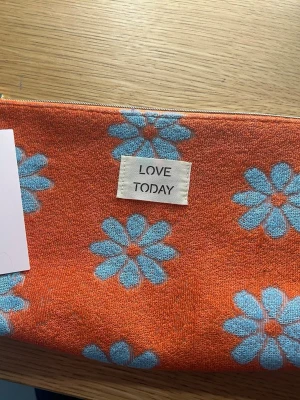 Orange necessär med blommor - Säljer en orange necessär med ljusblå blommor och texten 'LOVE TODAY' på framsidan. Väskan kommer från Nya Zeeland, har dragkedja upptill och är tillverkad i ett mjukt tygmaterial. Perfekt för småprylar eller smink.