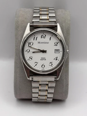 Klassisk silverfärgad klocka Moretime - Stilren klocka från Moretime med vit urtavla, svarta siffror och datumvisning. Armbandet är i silverfärgad metall med diskreta gulddetaljer. Klockan har en rund form och klassisk design som passar till många stilar.