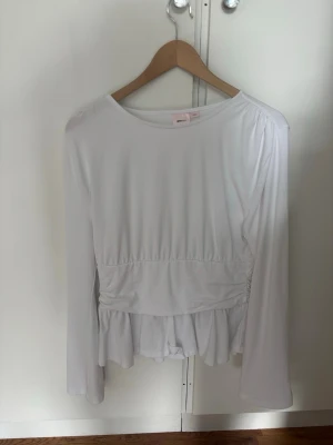 Vit långärmad topp med volang - Säljer en vit långärmad topp med rund hals och volangdetalj nedtill. Toppen har en markerad midja och är tillverkad i ett mjukt, stretchigt material. Perfekt för dig som gillar en feminin och enkel stil.
