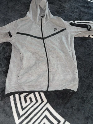 Grå zip-hoodie från Nike - Snygg grå hoodie från Nike med dragkedja framtill och svarta detaljer. Tröjan har huva, två sidofickor och en cool svart linje över bröstet. Perfekt för en sportig och avslappnad stil. Materialet är mjukt och bekvämt, troligen bomullsblandning.