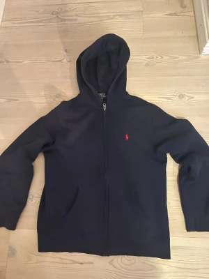 Mörkblå hoodie med dragkedja från Ralph lauren  - Snygg mörkblå hoodie från Polo Ralph Lauren med dragkedja framtill och klassisk röd logga broderad på bröstet. Hoodien har två fickor fram och en stor huva. Perfekt för en avslappnad och stilren look.