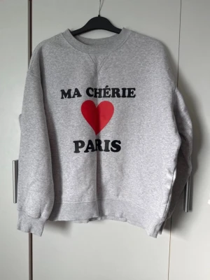 Grå sweatshirt med tryck - Grå sweatshirt med svart text 'MA CHÉRIE PARIS' och ett rött hjärta på bröstet. Tröjan har rund halsringning, ribbade muddar vid ärmslut och nederkant. Tillverkad i mjukt sweatshirtmaterial, perfekt för en avslappnad stil.