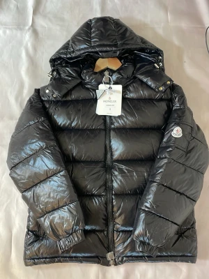 Moncler Maya - Snygg svart dunjacka från Moncler med glansig finish, stor luva och dragkedja framtill. Jackan har Moncler-logga på ärmen, två fickor med dragkedja och elastiska muddar vid ärmsluten. Perfekt för kalla vinterdagar och riktigt trendig look.