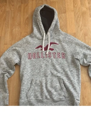 Grå Hollister hoodie med logga - Säljer en gråmelerad hoodie från Hollister med stor vinröd logga och text på bröstet. Hoodien har huva med vita snören och en stor magficka. Insidan är mjuk och fleecefodrad för extra komfort. Perfekt för chill dagar eller när du vill vara extra cozy.