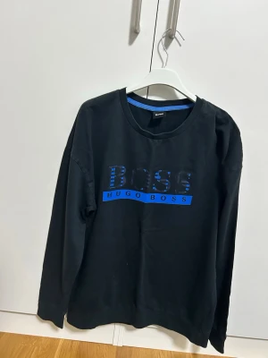 Svart långärmad tröja Hugo Boss storlek M - Svart långärmad tröja från Hugo Boss med blå logga och text på bröstet. Tröjan har rund halsringning och en avslappnad passform. Perfekt för en stilren och enkel look.