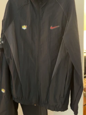 Svart Nike tracksuit med Tn-detaljer - Säljer en svart tracksuit från Nike med Tn-logga på både jacka och byxa. Jackan har dragkedja framtill, elastiska muddar och röd Nike Swoosh på bröstet. Byxorna har smal passform och matchande Tn-detaljer. Perfekt för träning eller chill.