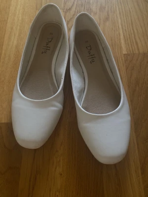 Vita ballerinaskor - Säljer ett par klassiska vita ballerinaskor från Duffy med rundad tå och låg klack. Skorna har en stilren design och är tillverkade i ett slätt material som ger en clean look. Perfekta att matcha med olika outfits. Använda ändats vid två tillfällen❤️❤️