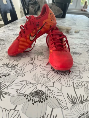 Röda Nike fotbollsskor med dobbar - Snygga röda fotbollsskor från Nike med orange och svarta detaljer samt vit Nike-logga på sidan. Skorna har snörning och plastdobbar för bra grepp på gräsplan. Tillverkade i syntetmaterial med grafiskt mönster på hälen och sidan. Perfekta för dig som vill sticka ut på planen.