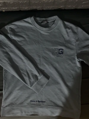 Ljusblå sweatshirt från Dolce & Gabbana - Snygg ljusblå sweatshirt från Dolce & Gabbana med DG-logga broderad på bröstet och texten 'Dolce & Gabbana' nedtill. Tröjan har rund halsringning och långa ärmar. Perfekt för en avslappnad och trendig look. De står ingen size på tröjan de jag kolla fast den passar som size M