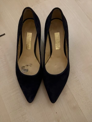 Svarta mocka-pumps från Gabor - Stilrena svarta pumps från Gabor i mjuk mocka med spetsig tå och låg klack. Klassisk och elegant design som passar till många olika looks. Insidan är beige och skon har en smal siluett för en snygg och tidlös känsla.