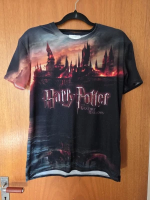 Harry Potter Deathly Hallows T-shirt - Cool t-shirt med tryck från Harry Potter and the Deathly Hallows. Motivet visar Hogwarts i mörka och röda toner, med texten på både fram- och baksidan. T-shirten har rund hals och korta ärmar, perfekt för alla Potterheads.
