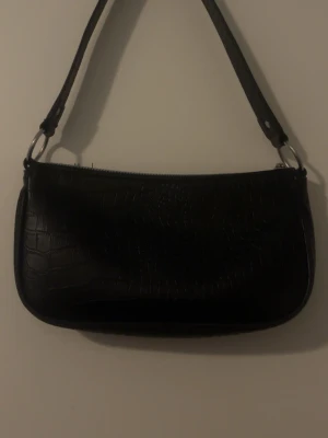Svart axelväska i krokopräglat material - Snygg svart handväska med axelrem och croco-mönstrad yta. Perfekt när man vill ha en stilren look men vill liva upp den lite med mönster. Köpt för några år sedan på HM, säljs då den inte kommer till användning. Har använt den ungefär 5ggr. De är bara att skriva vid frågor och priset kan såklart diskuteras👜