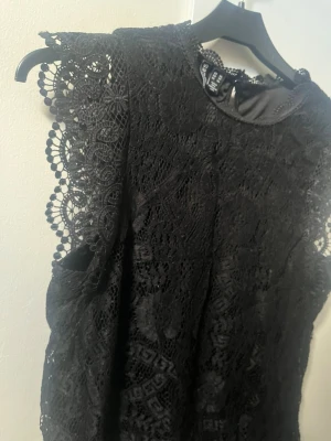 Svart spetsblus med detaljer - Säljer en svart blus i spets med fina broderade detaljer och ärmlös design.