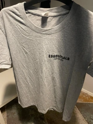 Grå Essentials t-shirt - Snygg grå t-shirt från Essentials med tryck på bröstet och en liten logga på ryggen. Klassisk rund halsringning och korta ärmar. Tillverkad i mjuk bomull som känns skön mot huden. Perfekt till en avslappnad stil.