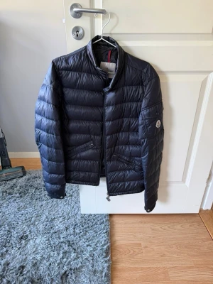 Mörk blå dunjacka från Moncler - Snygg mörkblå dunjacka från Moncler med quiltad design och ståkrage. Jackan har dragkedja framtill, två sidofickor och Moncler-logga på ärmen. Perfekt för kalla vinterdagar och riktigt stilren look. (Ny pris 10.000kr)