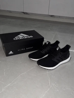 Adidas Ultra Boost svarta sneakers - Svarta Adidas Ultra Boost sneakers med stickad ovandel och vita Boost-sulor. Skorna har klassisk låg profil, svarta snören och diskreta Adidas-loggor på plösen och hälen. Perfekta för dig som gillar stilrena och bekväma sneakers.