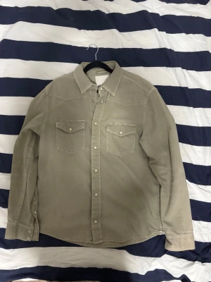 Beige manchesterskjorta med knappar - Snygg beige skjorta i manchester med klassisk krage och tryckknappar framtill. Två bröstfickor med lock och långa ärmar med knappslå. Perfekt för en avslappnad men ändå stilren look.