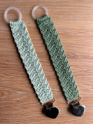 Gröna virkade napphållare med hjärta - Två handgjorda napphållare i ljusgrönt garn med virkade band och metallclips formade som hjärtan. Ena änden har en genomskinlig plastring för enkel fastsättning. Perfekt accessoar för att hålla koll på nappen med stil. 