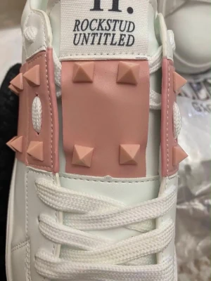 Valentino Rockstud Untitled sneakers - Snygga vita sneakers från Valentino med rosa detaljer och ikoniska pyramidnitar. Skorna har en chunky sula, vita skosnören och coola rosa paneler med nitar över ovandelen. Perfekta för dig som vill sticka ut med en lyxig och edgy stil.