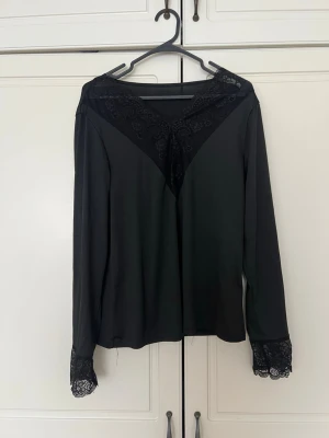 Svart blus med spetsdetaljer - Snygg svart blus med lång ärm och spetsdetaljer vid halsringningen och ärmsluten. Blusen har en V-formad spetsinfällning framtill och är tillverkad i ett mjukt, lätt material som ger en elegant look. Perfekt för dig som gillar stilrena plagg med en twist.